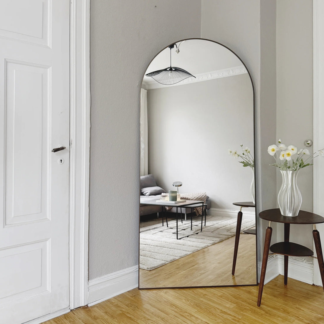 An arched floor mirror with a thin black frame stands in a bright hallway with light gray walls and wood floors, reflecting a living room with a sofa and coffee table. A white door is on the left, and a small wooden side table with a vase of flowers is on the right.